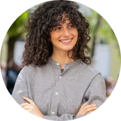 Portret van Anneliese Van Den Berg met krullend haar en gekruiste armen, glimlachend buiten.