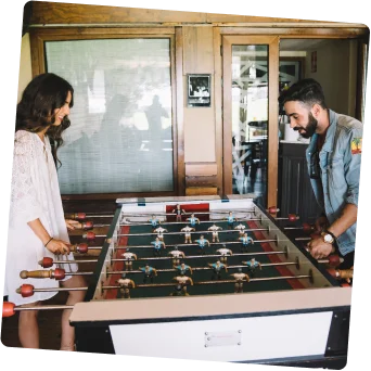 Drie mensen spelen vrolijk tafelvoetbal in een gezellige kamer met houten accenten. De scène is levendig en ongedwongen, met warme verlichting die de vriendelijke sfeer versterkt.