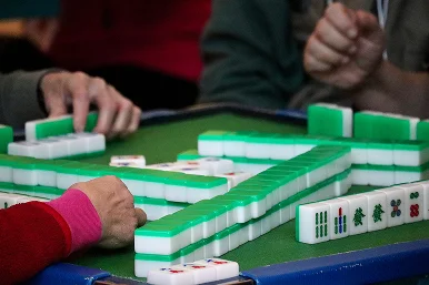 Drie mensen spelen Mahjong op een groene tafel, terwijl ze met hun handen tegels verplaatsen. De scène is gefocust en straalt concentratie en strategie uit.