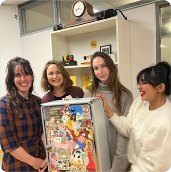 Vier vrouwen die samen glimlachend een creatieve projectpresentatie tonen met een kleurrijk bord vol illustraties.
